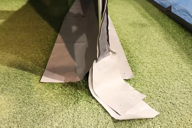 Nahaufnahme eines grauen Reißverschlusses an einer Stoffkante mit aufgeklappter Stofflasche mit mehreren Löchern auf grünem Kunstrasen; im rechten Hintergrund blaues Material