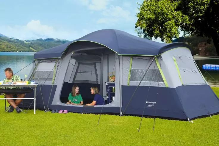 Großes blau-graues Zelt auf Gras am Ufer eines Sees; vorderer Eingang offen, zwei Personen sitzen im Eingangsbereich, eine Person sitzt links an einem Tisch; rechts ein Baum, im Hintergrund See, Hügel und teilweise bewölkter Himmel