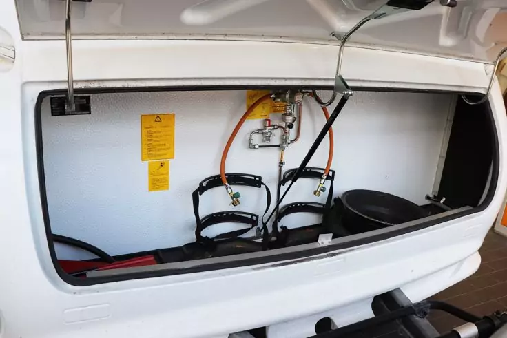 Offenes weißes Staufach mit zwei schwarzen Halterungen, orangefarbenem und schwarzem Schlauch, metallischer Armatur mit Anschlüssen, gelbem Aufkleber an der Innenwand, rundem schwarzen Deckel rechts sowie sichtbaren roten und grünen Kabeln