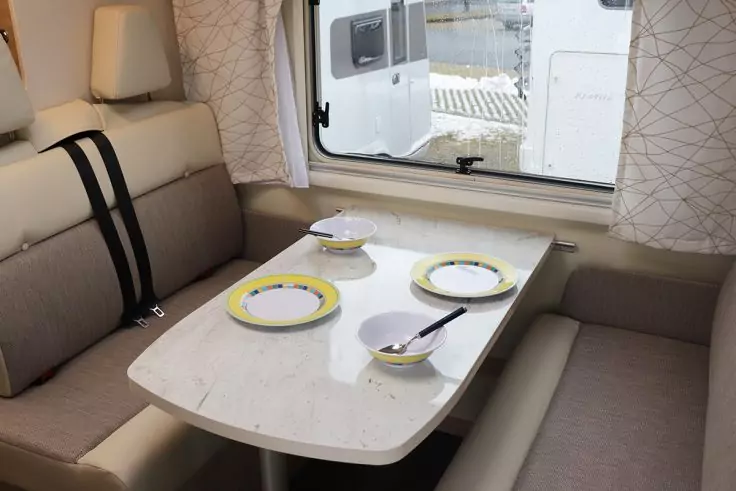 Essbereich eines Wohnmobils mit Tisch, vier Tellern mit gelbem Rand, vier Schalen mit Löffeln, Sitzbänken und Fenster mit Vorhang