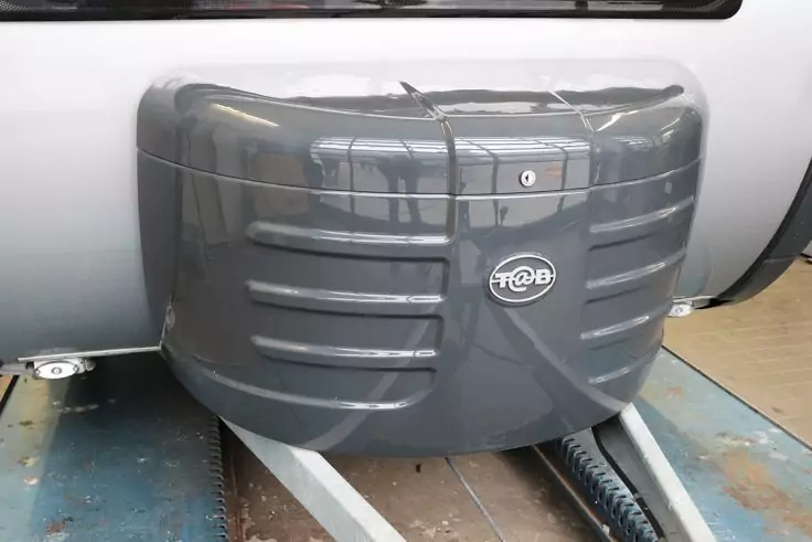 Dunkelgraue, abgerundete Kunststoffabdeckung mit horizontalen Rillen und rundem Emblem, montiert auf Metallschienen; schwarzer Kabelschlauch im Vordergrund, heller Fahrzeugkörper im Hintergrund