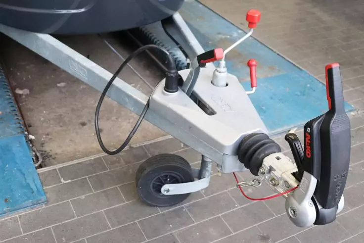 Nahaufnahme des vorderen Bereichs einer Metall-Anhängerkupplung mit Kupplungskopf, Handhebel, Sicherheitskabel und Stützrad auf gefliestem Boden