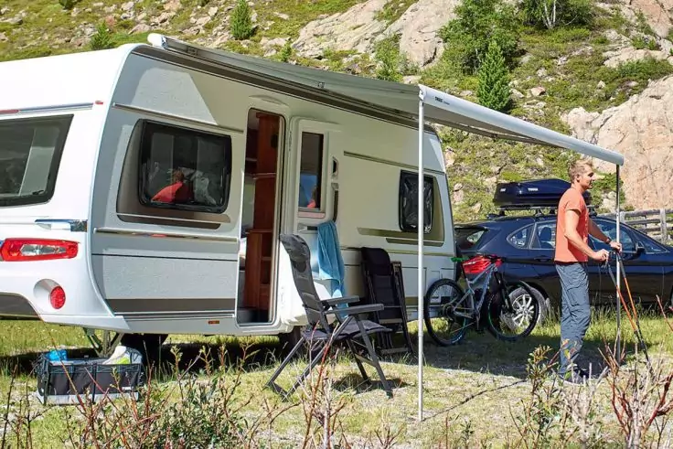 Weißer Wohnwagen mit geöffneter Seitentür und ausgefahrener Markise; zwei Klappstühle davor, ein Auto mit Fahrrad daneben, eine Person steht mit Wanderstöcken neben dem Auto; felsige, bewaldete Landschaft im Hintergrund