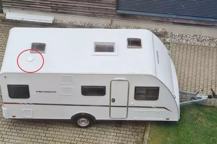 Weißer Wohnwagen auf gepflastertem Stellplatz neben einem Gebäude, Draufsicht; drei Dachöffnungen und ein rundes weißes Objekt auf dem Dach, von einem roten Kreis markiert; Tür, Fenster, Rad und Anhängerkupplung seitlich sichtbar