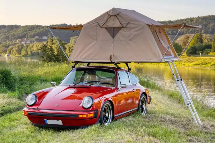 Roter klassischer Sportwagen mit aufgeklapptem beigefarbenem Dachzelt und einer Leiter zum Gras, geparkt neben einem See, Bäume und Hügel im Hintergrund