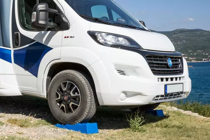 Vorderer linker Teil eines weißen Wohnmobils auf Gras an der Küste, blaue Unterlegkeile vor dem Vorderrad, Meer und Berge im Hintergrund
