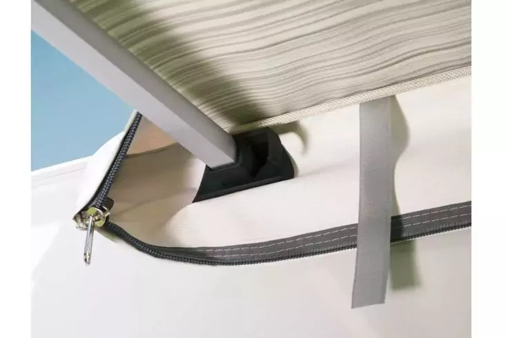 Nahaufnahme eines schwarzen Reißverschlusses an weißem Stoff mit Metallzipper, grauem Stoffriemen und schwarzem Kunststoffhalter; darüber grünes, gestreiftes Stoffpaneel und ein Stück blauer Himmel