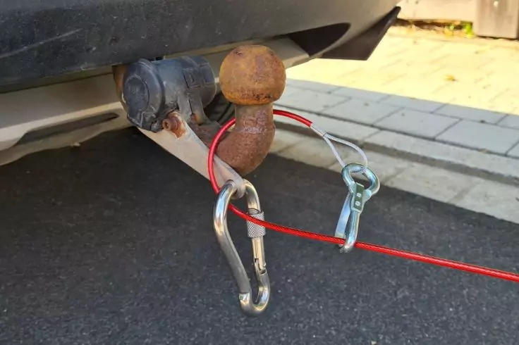 Heckstoßstange eines Fahrzeugs mit Anhängerkupplung; rostige Anhängerkugel; rotes Kabel mit Karabinerhaken und Metallklemme; Asphalt und Gehweg im Hintergrund