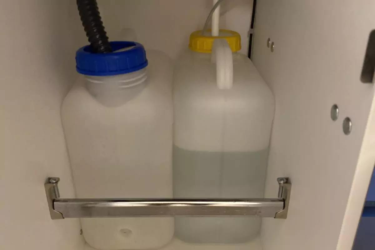 Zwei durchsichtige Kunststoffkanister in einem wei&szlig;en Schrank; links mit blauem Schraubverschluss und schwarzem Schlauch, rechts mit gelbem Verschluss und wei&szlig;em Griff, vorn eine Metallstange