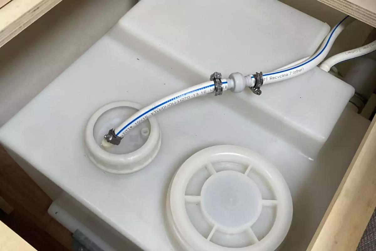 Wei&szlig;er Kunststoffbeh&auml;lter mit zwei runden Verschl&uuml;ssen und zwei angeschlossenen wei&szlig;en Schl&auml;uchen mit Metallschellen in einem Holzrahmen