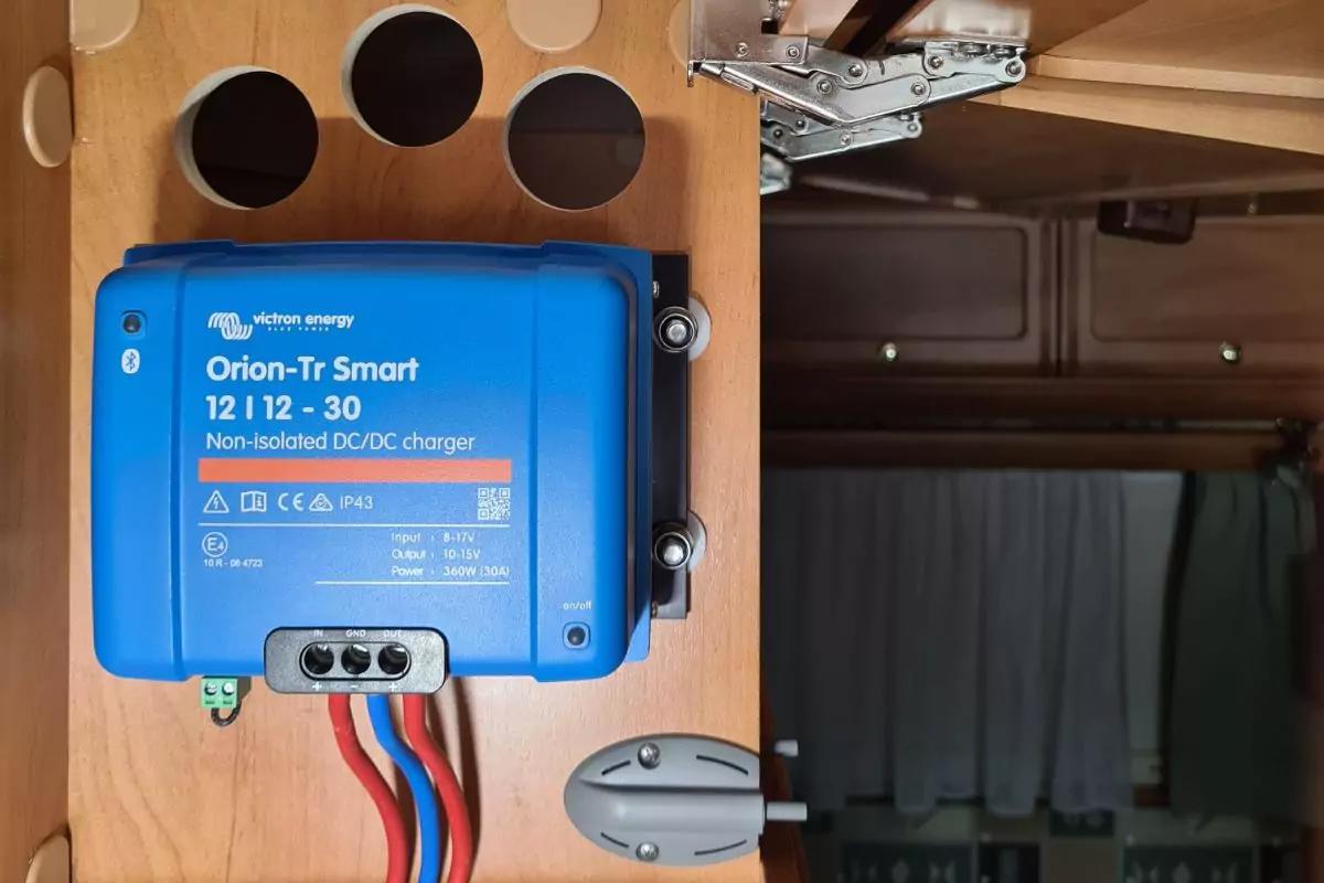 Blauer Kasten mit wei&szlig;er Aufschrift 'Orion-Tr Smart 12 | 12-30 Non-isolated DCDC charger', an einer Holzplatte montiert; drei runde Ausschnitte oberhalb; mehrere isolierte Kabel (rot, schwarz, blau) f&uuml;hren unten heraus; rechts ein Metallscharnier und