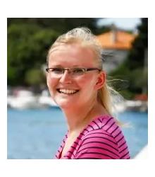 Person mit hellen, zur&uuml;ckgebundenen Haaren, Brille und pink gestreiftem Shirt, l&auml;chelnd vor Wasser und unscharfen Booten