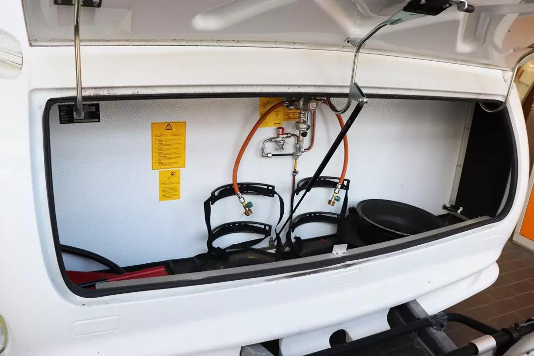 Offenes wei&szlig;es Abteil mit aufklappbarer Klappe; darin zwei orange und schwarze Schl&auml;uche an Metallarmaturen, zwei schwarze Haltegurte, ein gelbes Warnschild und eine schwarze runde Schale