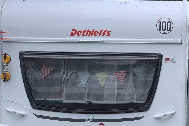 Wei&szlig;e Wohnwagenfront mit rotem Dethleffs-Logo, gro&szlig;em schwarz umrandetem Fenster, Wimpelkette und Gardine im Fenster, zwei gelbe runde Leuchten links, runde Plakette '100' oben rechts
