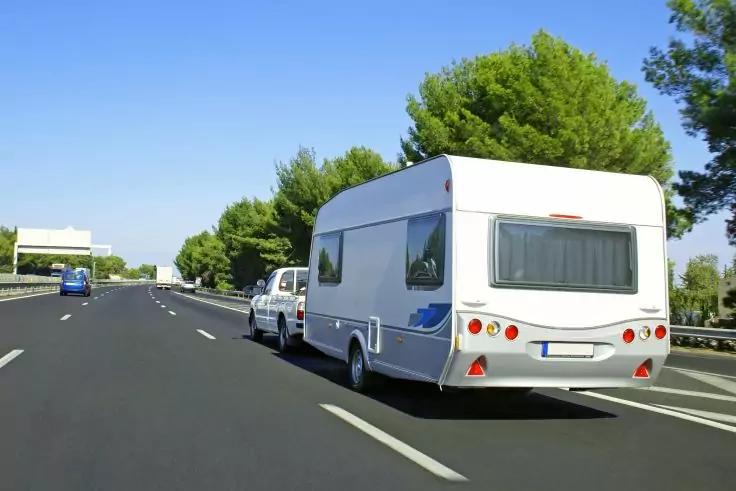 Wei&szlig;er Wohnwagen, an wei&szlig;em Pkw gekoppelt, auf mehrspuriger Autobahn mit Fahrbahnmarkierungen, B&auml;ume am Stra&szlig;enrand, blauer Himmel