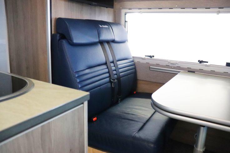 Zwei dunkelblaue gepolsterte Sitze mit Sicherheitsgurten neben einem Tisch und gro&szlig;em Fenster, helle Holzverkleidung und Arbeitsplatte links