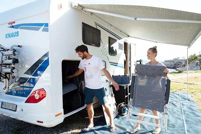 Wei&szlig;es Wohnmobil mit ausgefahrener Markise; ein Mann &ouml;ffnet ein seitliches Staufach, eine Frau tr&auml;gt zwei zusammengeklappte Klappst&uuml;hle auf einer blauen Plane; im Hintergrund Felsen und Wasser