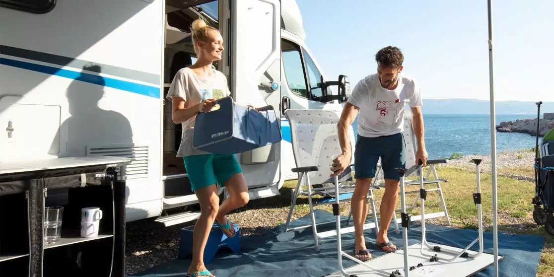 Zwei Erwachsene neben einem geparkten Wohnmobil an der K&uuml;ste; eine Person tr&auml;gt eine Sch&uuml;ssel und eine Box, die andere baut ein klappbares Tischgestell auf; blauer Bodenbelag, Meer und Himmel im Hintergrund