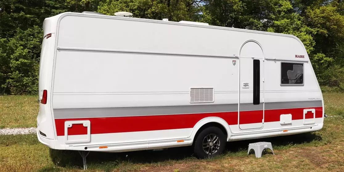 Wei&szlig;er Wohnwagen mit rotem Streifen, T&uuml;r und Fenster, auf Gras vor B&auml;umen geparkt