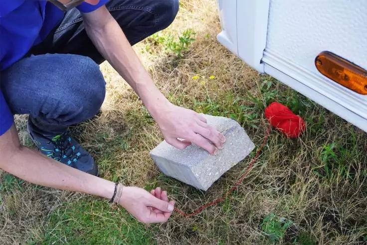 Person kniet auf Gras und legt einen grauen Betonblock neben die wei&szlig;e Seitenwand eines Wohnwagens; roter Gegenstand und orangefarbener Reflektor sichtbar