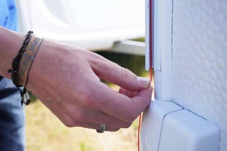 Nahaufnahme einer linken Hand mit Armb&auml;ndern und Ring, die einen d&uuml;nnen Dichtungsstreifen an den wei&szlig;en T&uuml;rrahmen eines Fahrzeugs h&auml;lt; unscharfer Grasboden im Hintergrund
