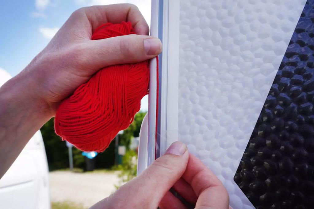 Zwei H&auml;nde halten ein Kn&auml;uel rote Wolle und einen wei&szlig;en Streifen an eine wei&szlig;e, strukturierte Fl&auml;che mit schwarzem Rechteck; unscharfer Hintergrund mit Auto und B&auml;umen