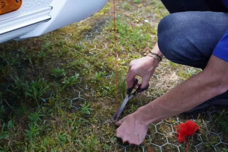 Person kniet auf Gras neben einer Auto-Sto&szlig;stange und schneidet mit einer Schere Drahtgitter; ein roter Gegenstand liegt auf dem Gitter