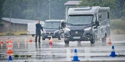 Zwei Wohnmobile fahren auf nasser Fahrbahn zwischen orange und blauen H&uuml;tchen; links steht eine Person in dunkler Kleidung; Geb&auml;ude und B&auml;ume im Hintergrund