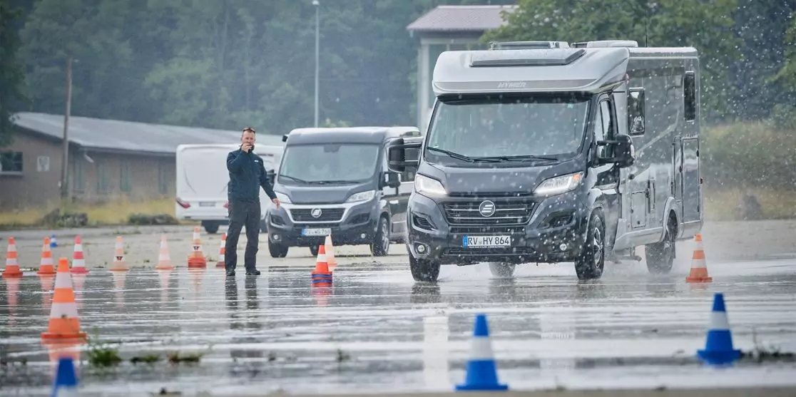 Zwei Wohnmobile fahren auf nasser Fahrbahn zwischen orange und blauen H&uuml;tchen; links steht eine Person in dunkler Kleidung; Geb&auml;ude und B&auml;ume im Hintergrund
