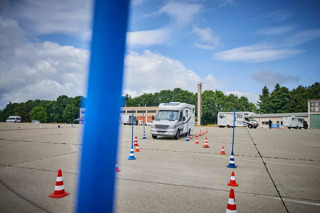 Gro&szlig;es befestigtes Areal mit orangen Verkehrsh&uuml;tchen und blauen Pfosten, Wohnmobil in der Mitte, Geb&auml;ude, B&auml;ume und bew&ouml;lkter Himmel im Hintergrund
