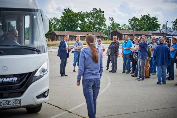Wei&szlig;er Bus links, Person mit Pferdeschwanz geht auf eine Gruppe von Menschen auf einem betonierten Platz zu; im Hintergrund B&auml;ume und niedrige Geb&auml;ude, bew&ouml;lkter Himmel