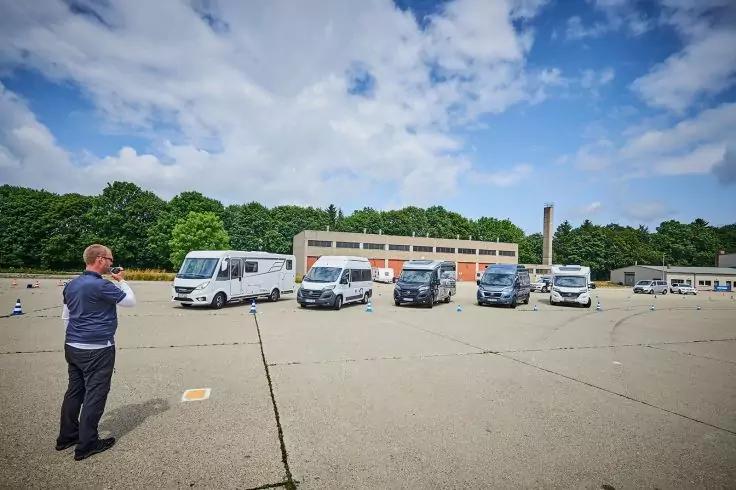 Person links mit Smartphone in der Hand vor mehreren in einer Reihe geparkten Transportern auf einer gro&szlig;en betonierten Fl&auml;che, im Hintergrund ein Geb&auml;ude, B&auml;ume und bew&ouml;lkter Himmel