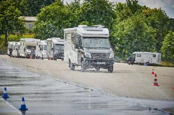 Mehrere wei&szlig;e Wohnmobile fahren auf einer nassen Fl&auml;che, das vordere Fahrzeug spritzt Wasser, rechts stehen Verkehrskegel, im Hintergrund B&auml;ume