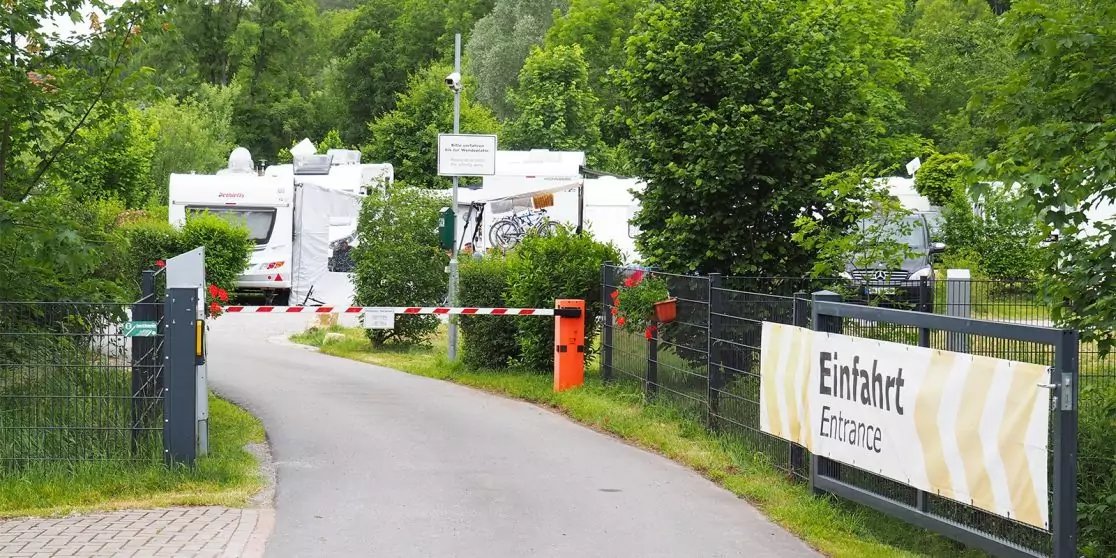 Zufahrtsstra&szlig;e mit Schranke, orangener Kassenautomat, mehrere Wohnmobile im Hintergrund, Schild 'Einfahrt Entrance', Zaun und B&auml;ume