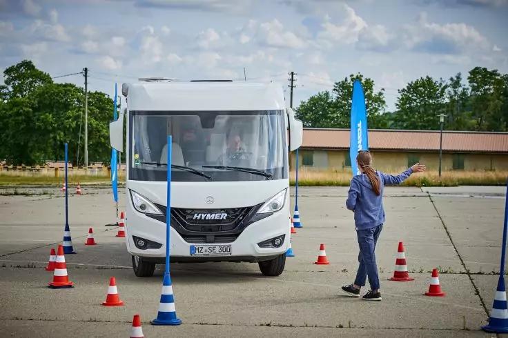 Wei&szlig;es Wohnmobil von vorn auf einem Asphaltplatz mit rot-wei&szlig;en Pylonen und blauen Pfosten; eine Person in blauer Kleidung h&auml;lt einen blauen Pfosten neben dem Fahrzeug; niedriges Geb&auml;ude, B&auml;ume und bew&ouml;lkter Himmel im Hintergrund