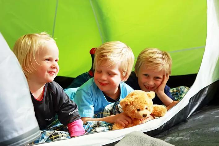 Drei Kinder liegen in einem Zelt auf Schlafs&auml;cken, eines h&auml;lt einen Teddyb&auml;r