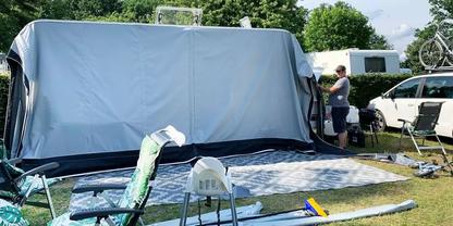 Gro&szlig;e graue Vorzeltfront vor einem Wohnwagen. Vor dem Vorzelt liegen eine gemusterte Bodenmatte, Campingst&uuml;hle und Campingutensilien auf einer Wiese. Rechts steht ein Auto mit Fahrrad auf dem Dach; zwei Personen stehen neben dem Vorzelt. B&auml;ume und wo