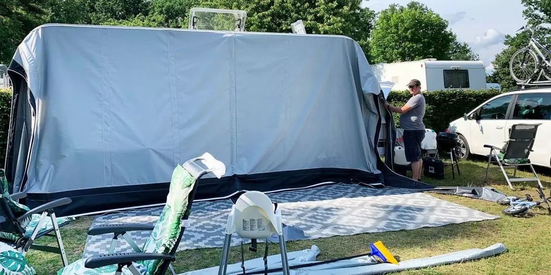 Gro&szlig;e graue Vorzeltfront vor einem Wohnwagen. Vor dem Vorzelt liegen eine gemusterte Bodenmatte, Campingst&uuml;hle und Campingutensilien auf einer Wiese. Rechts steht ein Auto mit Fahrrad auf dem Dach; zwei Personen stehen neben dem Vorzelt. B&auml;ume und wo