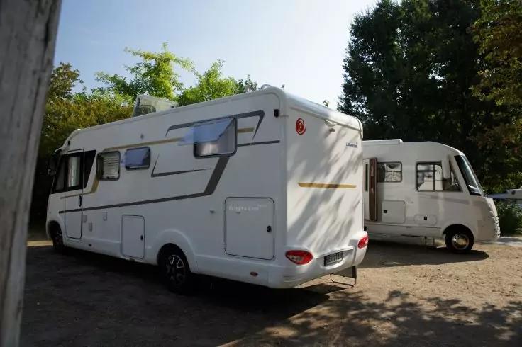 Zwei wei&szlig;e Wohnmobile nebeneinander; vorne das Heck eines Wohnmobils, dahinter die Seite eines weiteren; B&auml;ume und Schatten