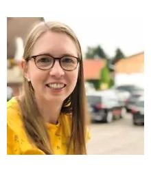 Frau mit Brille und langen blonden Haaren, gelbes Oberteil, l&auml;chelnd im Freien vor geparkten Autos und Geb&auml;uden im unscharfen Hintergrund