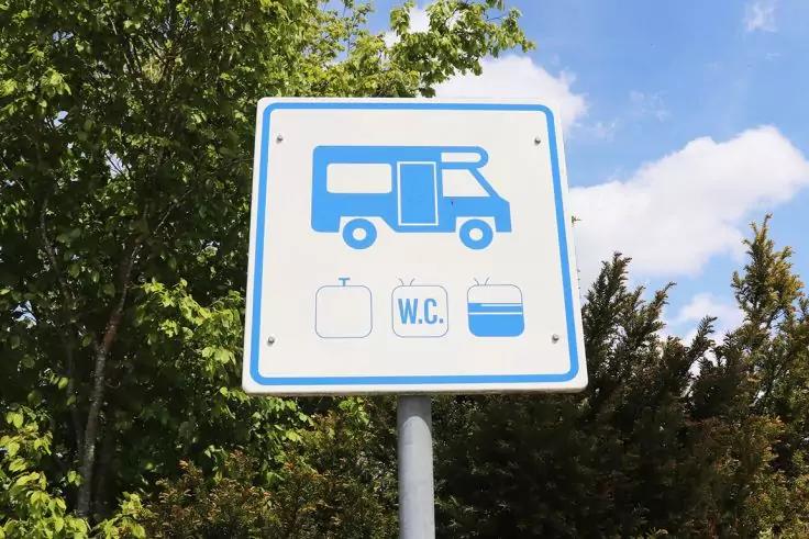 Quadratisches wei&szlig;es Schild mit blauem Rand auf Metallpfosten vor B&auml;umen und Himmel; oben blaues Piktogramm eines Wohnmobils, darunter drei blaue Symbole: Gasflasche, Schriftzug &sbquo;W.C.&lsquo; und rechteckiger Beh&auml;lter