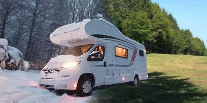 Wei&szlig;es Wohnmobil mit Aufbau &uuml;ber der Fahrerkabine und eingeschalteten Lichtern, geparkt mit Schnee links und gr&uuml;nem Gras rechts, beleuchtete Fenster