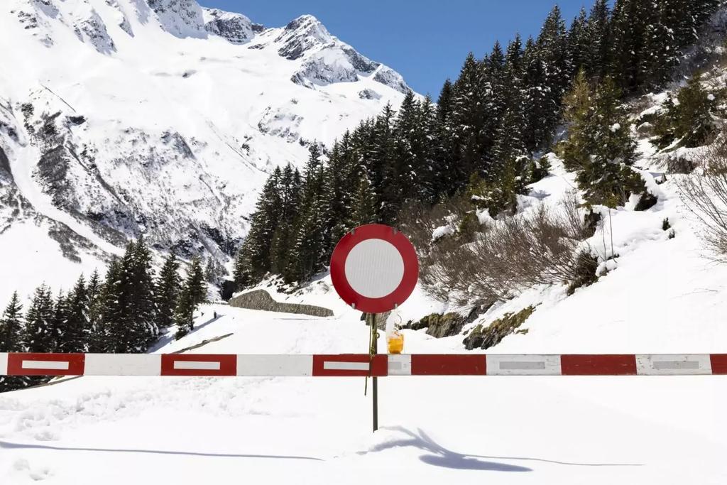 Schneebedeckte Stra&szlig;e blockiert von rotem-wei&szlig;en Schrankenbalken und rundem Schild mit rotem Rand und wei&szlig;er Mitte; im Hintergrund verschneite Tannen und Berggipfel unter blauem Himmel