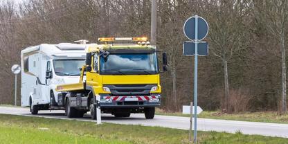 Gelber Abschleppwagen mit aufgefahrenem wei&szlig;em Wohnmobil auf einer Landstra&szlig;e, Verkehrsschilder, Leitplanke und B&auml;ume im Hintergrund