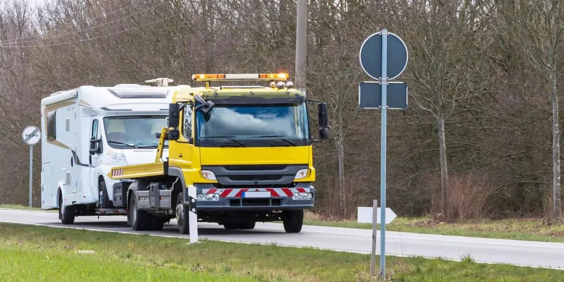 Gelber Abschleppwagen mit aufgefahrenem wei&szlig;em Wohnmobil auf einer Landstra&szlig;e, Verkehrsschilder, Leitplanke und B&auml;ume im Hintergrund