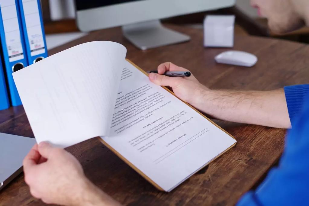 Person h&auml;lt Klemmbrett mit bedruckten Seiten &uuml;ber einem Holztisch; H&auml;nde, Kugelschreiber, Monitor, Maus und Aktenordner sichtbar