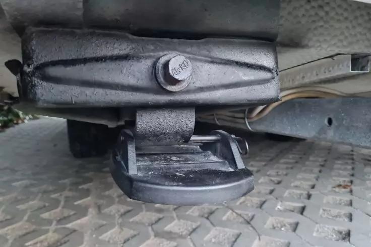 Unterseite eines Fahrzeugs mit schwarzem Metallbauteil, gro&szlig;er Schraube oben und rundem flachen Teil darunter; gepr&auml;gte Metallplatte im Vordergrund