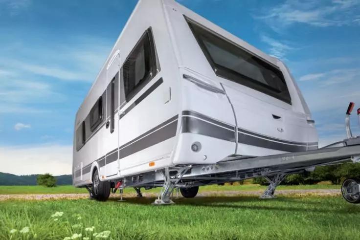 Wei&szlig;er Wohnwagen auf Wiese, Vorderseite mit Deichsel, St&uuml;tzbeinen und Fenstern, blauer Himmel im Hintergrund