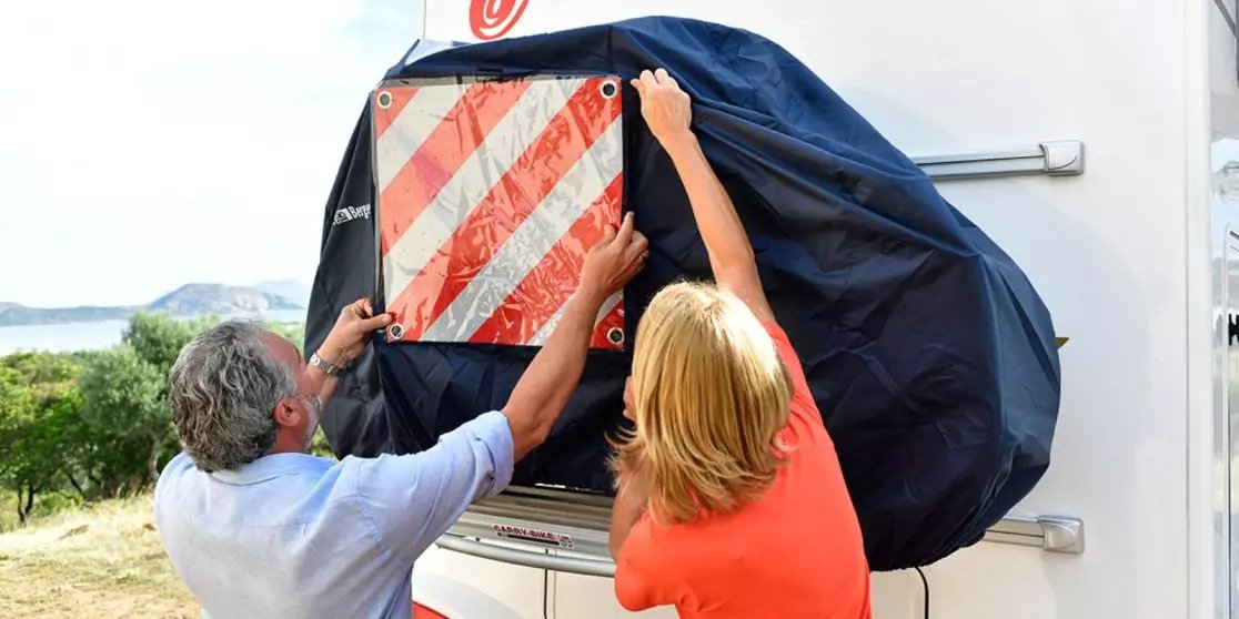 Zwei Personen (Mann und Frau) befestigen ein rot-wei&szlig; gestreiftes Warnschild an der R&uuml;ckseite eines wei&szlig;en Wohnwagens, schwarzer Schutzbezug sichtbar, im Hintergrund Wasser und H&uuml;gel