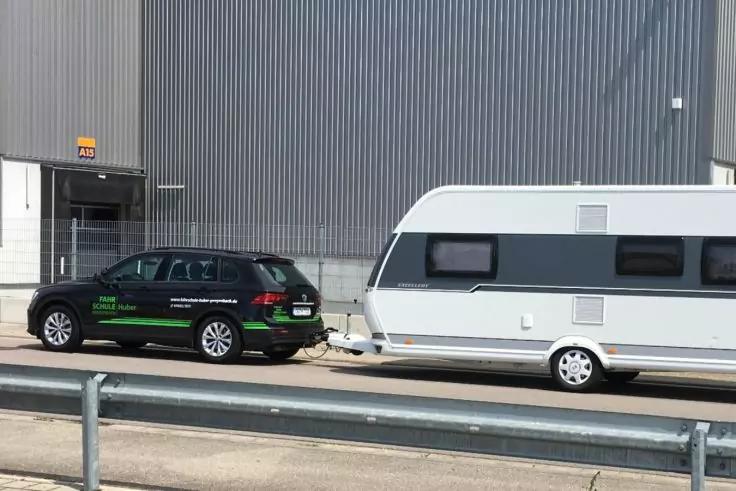 Schwarzer Kombi mit gr&uuml;ner Beschriftung, angekoppelter wei&szlig;er Wohnwagen vor grauer Wellblechfassade, Metallgel&auml;nder im Vordergrund